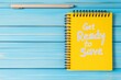 © Eugen - On a blue wooden table, there lies a cheerful yellow notebook featuring the motivational phrase get ready to save written on its cover, sitting next to a sharpened pencil