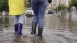 © TO LOVE - girls in raincoats and rubber boots walk along road flooded with torrential rains, their feet walk through puddles city, splashing water to the sides, the flood is on street, car is driving on water.