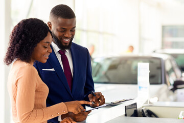 Wall Mural - Happy Black Woman Discussing Agreement Details With Manager While Buying Car In Showroom, Young Smiling African American Female Choosing New Automobile In Modern Dealership Center, Free Space