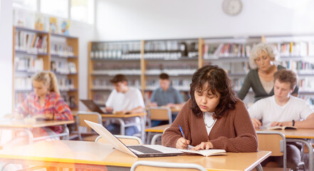 Wall Mural - Fifteen-year-old Asian schoolgirl is studying in the school library on a laptop while preparing for an exam, making important notes in a copybook