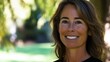© JO BLA CO - A cheerful woman stands outdoors in a park, smiling warmly at the camera. Bright sunlight filters through the trees, enhancing the vibrant green background