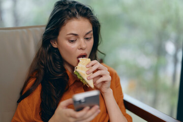 Canvas Print - Young woman enjoying a sandwich while using her smartphone in a cozy setting, showcasing a modern lifestyle and healthy eating habits