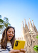 © CarlosBarquero - Vertical. Young women friend using mobile phone in hands in front of Sagrada Familia in Barcelona. Beautiful caucasian girls at European city enjoying vacation happy and smiling. Funny tourism people