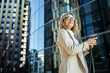 © muse studio - Businesswoman using smartphone while standing outside modern office building on sunny day
