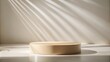 © sasiprapa - Cream-colored cylindrical display platform illuminated by sun rays casting shadows on a light beige wall and floor.