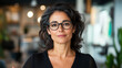 © Arty&Happy - woman with dark hair wearing glasses looks forward calmly and professionally, inside an office space