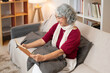 © bongkarn - A happy Asian senior retired woman enjoys writing in her dairy on a sofa in the living room.