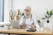 © sofiko14 - Young Muslim female doctor in hijab has online consultation with patient, writing notes during video call. Modern medical professional working remotely, enhancing patient communication.