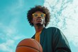 © Irina Kozel - A stylish young man with an afro hairstyle holds a basketball, wearing yellow glasses against a bright blue sky with clouds