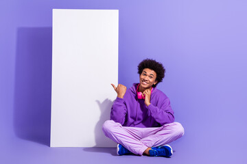 Full length photo of optimistic nice guy sitting indicating at smartphone screen touching headphones isolated on violet color background
