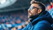 © khonkangrua - Thoughtful man in glasses enjoys live sporting event while sitting in stadium with blurred crowd in background