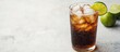 © CreativeBro - Refreshing dark soda drink served in a clear glass with ice and lime wedge on a light gray table with fresh limes in the background and empty space for text