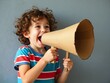 © dibas99 - A young boy with curly hair stands in front of a gray wall, excitedly using a cardboard megaphone. He wears a colorful striped shirt and radiates joy while pretending to communicate loudly.