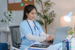 © amnaj - Asian female doctor wearing blue uniform and stethoscope is working with laptop at desk in clinic office