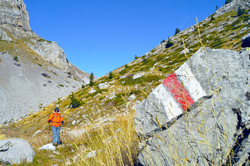 Wall Mural - Hiking routes in the mountains of Albania: a close-up photograph of a rock with red and white tourist markings and a hiker walking along the trail in the background. Male tourist in Buni i Jezerces