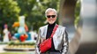 © Prana - Stylish older man at a modern outdoor sculpture garden, wearing a sleek silver jacket and bold red bag (Travel) (Aging)