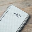 © Tanakorn - A notebook with a 'weight loss plan' written on the cover, resting on a wooden surface, symbolizing health and fitness planning.
