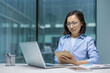 © Liubomir - Serious and focused Asian woman listening to online webinar, remote business meeting conference. Woman in the middle of the office working with laptop, writing data in notebook.