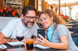 © simona - A couple of 48 years old enjoys a leisurely lunch outside at a restaurant, laughing together as the woman shares funny mobile content with her husband, in a peaceful outdoor setting under the sun.
