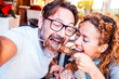 © simona - Two old friends seated at a restaurant table, enjoying a grilled meat skewer while smiling and laughing together. One of them is wearing glasses and the other has curly blonde hair, sharing a selfie.