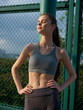 © SHOTPRIME STUDIO - Young woman in athletic wear posing confidently outdoors, basking in sunlight against a green fence backdrop