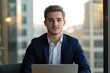 © Roshani - Young businessman in a suit sits before a laptop, looking directly at the camera in a modern office setting.