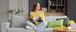 © Pixel-Shot - Young woman with magazine sitting on grey sofa in living room