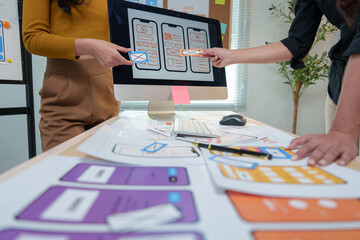 Two ux designers are comparing mobile application wireframe sketches on a desktop computer, analyzing different layouts and user interface elements for optimal user experience