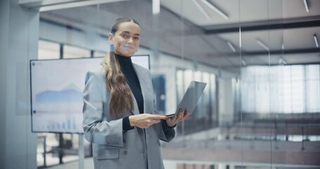 Aufkleber - Smiling Caucasian Woman Connecting with Clients Online on a Laptop. Portrait of a Young Female Manager Standing in Office Setting in a Grey Jacket, Looking at Camera and Holding a Computer