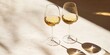 © Lala - Two elegant glasses of white wine on a light beige surface with soft shadows and bright highlights reflecting natural sunlight during the day