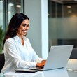 © hs - Una ejecutiva hispana trabaja con su computadora portátil en su oficina. Está sonriendo.