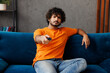 © ViDi Studio - Young frustrated bored Indian man wear orange casual clothes sits on blue sofa couch stay at home switch channel watch tv hotel flat rest relax spend free time in living room indoor. Lounge concept.