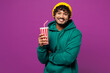 © ViDi Studio - Young happy smiling fun Indian man he wear green hoody yellow hat casual clothes hold in hand cup of soda pop cola fizzy water isolated on plain purple background studio portrait. Lifestyle concept.