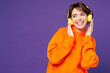 © ViDi Studio - Young smiling happy Caucasian woman she wear orange hoody casual clothes listen to music in headphones look aside on area mockup isolated on plain purple background studio portrait. Lifestyle concept.