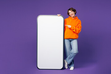 Wall Mural - Full body young woman she wears orange hoody casual clothes point index finger on big huge blank screen mobile cell phone smartphone with area isolated on plain purple background. Lifestyle concept.