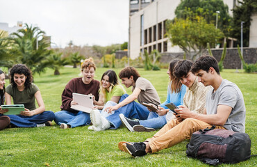 Happy students studying with digital tablets sitting on grass at campus park with college building in background - Back to school concept - Main focus on right boy face