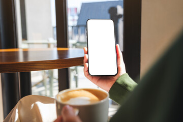 Wall Mural - Mockup image of a woman holding mobile phone with blank desktop screen while drinking coffee in cafe