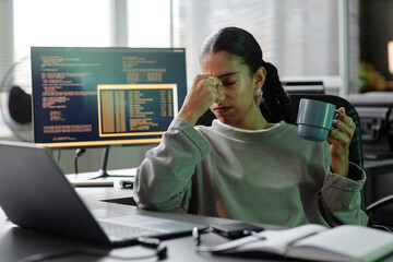 Medium shot of overworked female programmer sitting at desk with cup of coffee touching tired eyes at office workplace, copy space