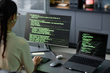 Over shoulder shot of female IT programmer typing source codes on computer using multiple screens working at office workplace