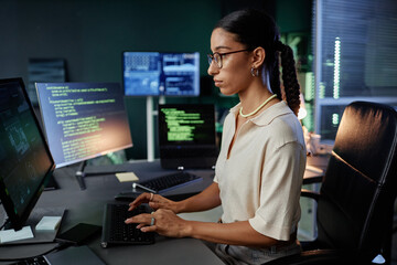 Side view of Middle Eastern female IT programmer immersed in typing on computer at workstation working in evening hours in empty office, captured against multiple screens with lines of code running