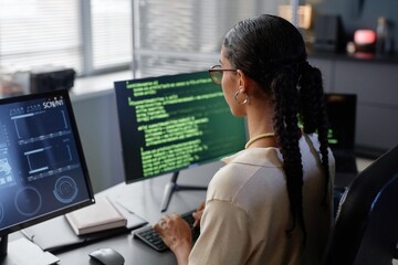 Rear view of Middle Eastern female programmer with glasses reading multiple computer screens while working on code at professional workstation in IT company office