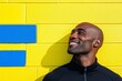 © PaulShlykov - Smiling man in black clothing leaning against a bright yellow wall with blue accents during daytime