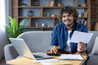 © Liubomir - Portrait of a young man at home doing paperwork, paying credit checks, utility bills, bank bills. Freelancer smiling happily and looking at camera sitting on sofa, using laptop.