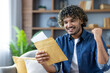 © Liubomir - Successful and joyful man sitting on sofa in living room, holding postal envelope letter in hands, celebrating victory and successful results obtained in notified .