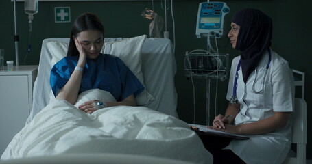 Wall Mural - Young African doctor sits next to a patient in a hospital room, examines her, asks about her health, and writes notes on paper about her health.