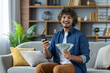 © Liubomir - Young successful man sitting at home on the couch holding phone and money cash American dollars in hands. Freelancer smiling and looking at camera, celebrating winning online app.