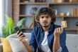 © Liubomir - Disappointed and deceived man looking at camera, young freelancer sitting at home on sofa in living room, holding bank credit card and phone, received account blocking, and money transfer error.