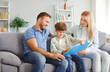 © Studio Romantic - Smiling parents sitting on couch with their cheerful son between them writing in notebook. Happy mother and father spending leisure time with child, helping him with homework. Family bonds concept.