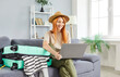 © Studio Romantic - Happy smiling woman traveler sitting on sofa at home with suitcase and booking tickets online via laptop. Young girl wearing beach hat getting ready for holiday trip. Vacation concept.