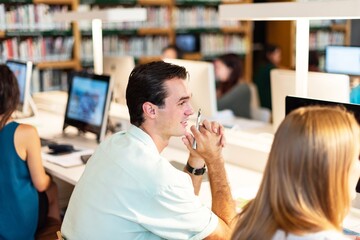 Poster - Man in white shirt sitting at a desk in a library. Focused man sitting in library with other study in the background. Caucasian man and other studying with computer in library. Happy man in library
